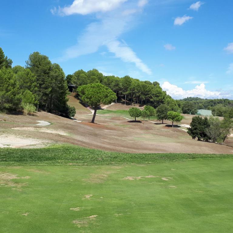 Panoramaaufnahme eines Golfplatzes mit grüner Rasenfläche, Sandbunkern und dichtem Baumbestand unter blauem Himmel.