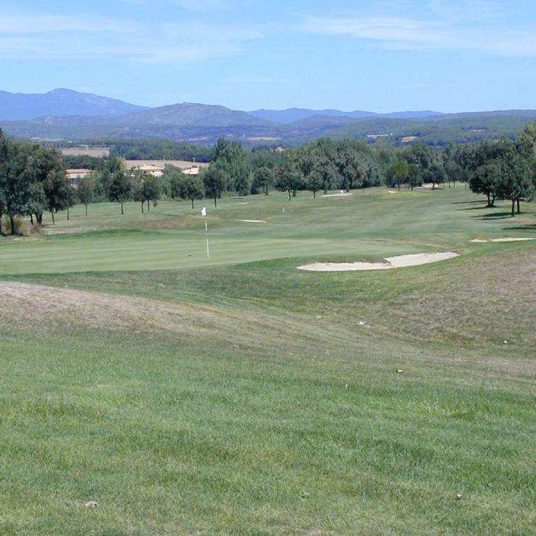 Ein weitläufiger Golfplatz mit Sandbunkern, Bäumen und Blick auf eine Bergkette unter blauem Himmel.