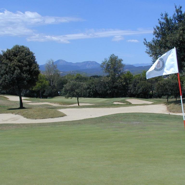 Panoramaaufnahme eines Golfplatzes mit grünem Rasen, einem Flaggenmast und einer Bergkulisse im Hintergrund.