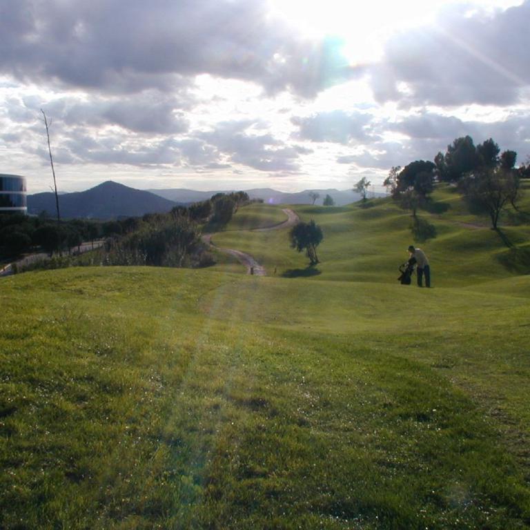 Eine weite, grüne Wiese mit sanft ansteigendem Gelände, das in eine Berglandschaft übergeht, unter einem dramatisch bewölkten Himmel.