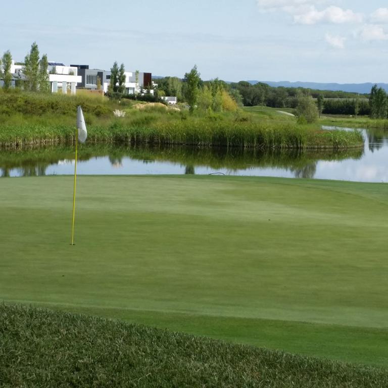Ein Golfplatz mit einem Wasserhindernis und modernen Gebäuden im Hintergrund.