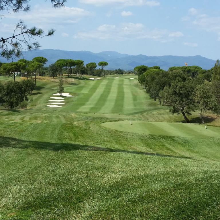 Weitwinkelaufnahme eines grünen Golfplatzes mit Bäumen und Bergpanorama unter blauem Himmel.