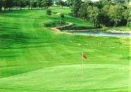 Ein weite Aufnahme eines grünen Golfplatzes mit einem Fahnenmast im Vordergrund, einem Wasserloch und Bäumen im Hintergrund.