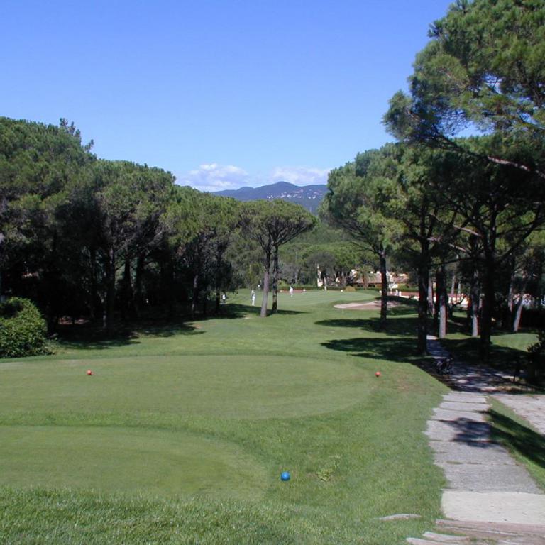 Ein Golfplatz mit einem großen Grün im Vordergrund, einem Weg, der zwischen hohen Pinien Bäumen verläuft, und einem Blick auf die ferne Bergkette unte