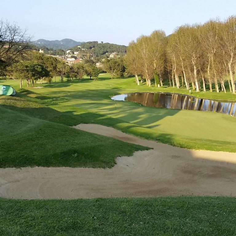 Ein Golfplatz mit Sandbunker, grünem Rasen, einem Teich und einer Reihe von Bäumen im Hintergrund.