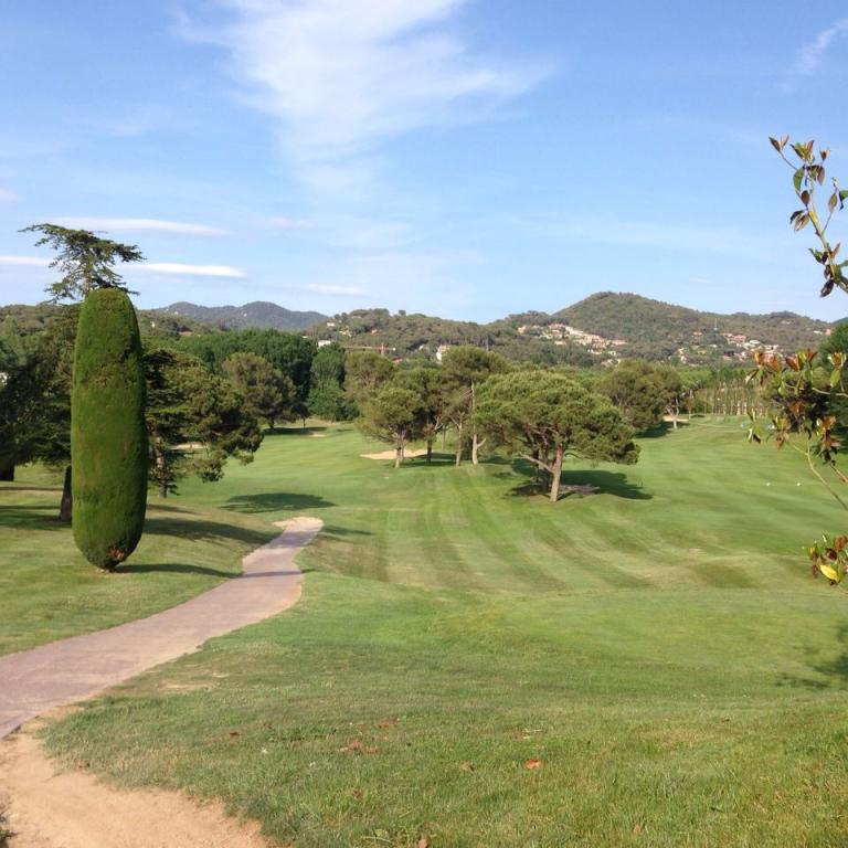 Panoramablick auf einen gepflegten Golfplatz mit einem geschwungenen Weg und bewaldeten Hügeln im Hintergrund.