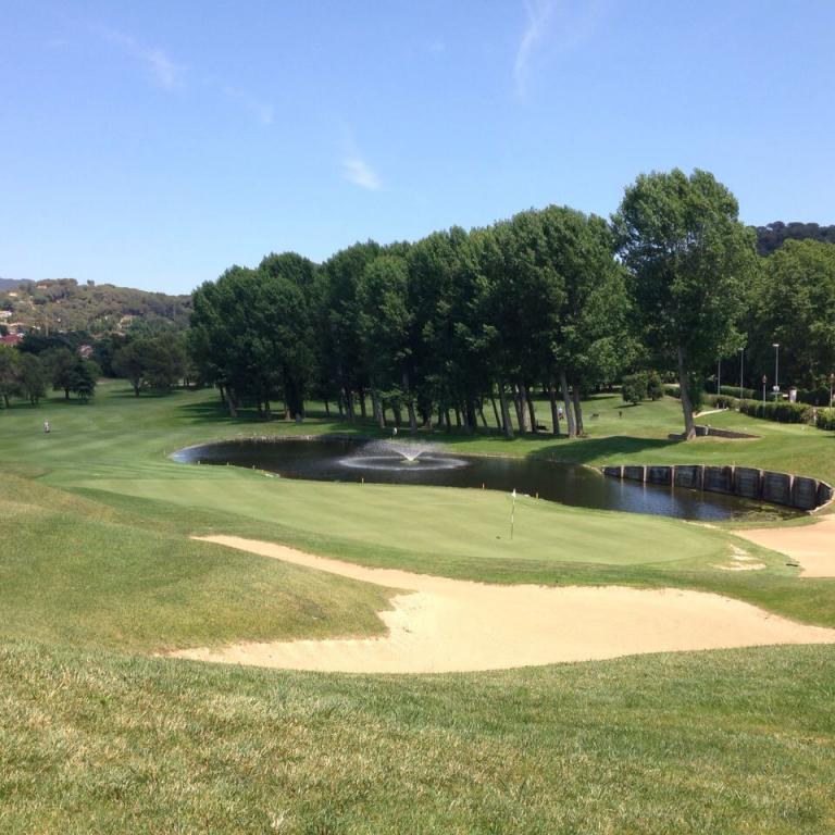 Weitwinkelaufnahme eines Golfplatzes mit Wasserbecken, Sandbunkern und einer Reihe von Bäumen unter blauem Himmel.