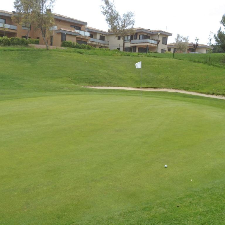 Ein weitläufiger, gepflegter Rasen eines Golfplatzes mit einem Fahnenmasten und luxuriösen Gebäuden im Hintergrund.