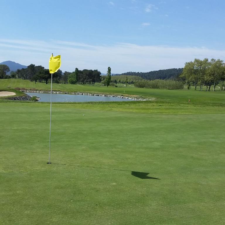 Weitwinkelaufnahme eines Golfplatzes mit gepflegtem Grün, einem Wasserbecken und bewaldeten Hügeln im Hintergrund.
