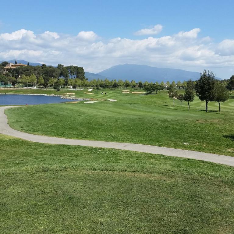 Ein weitläufiger, gepflegter Golfplatz mit einem geschwungenen Weg, einem Teich und Blick auf die bewaldeten Hügel im Hintergrund.