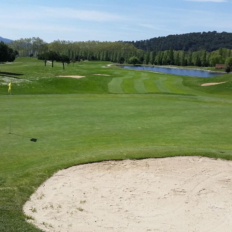 Weitwinkelaufnahme eines Golfplatzes, die einen großen Sandbunker im Vordergrund, einen gepflegten Rasen und im Mittelgrund ein Wasserbecken zeigt.