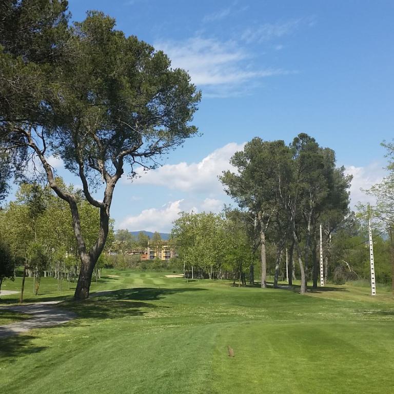 Ein weitläufiger, gepflegter Golfplatz unter einem blauen Himmel, gesäumt von großen Bäumen und einer klaren Wiese.