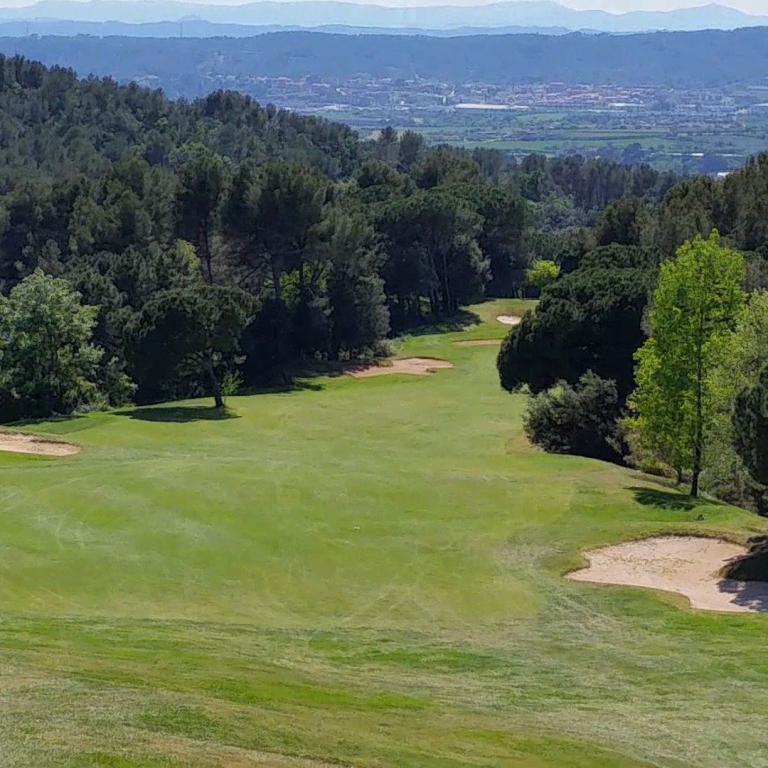 Ein Panoramablick auf einen Golfplatz mit grüner Wiese, Sandbunkern und einer dahinterliegenden Wald- und Stadtkulisse.