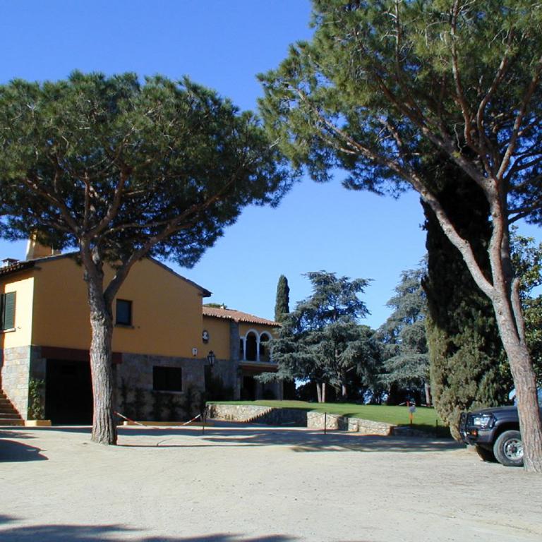 Eine weitläufige, gelbe Villa mit einem gepflasterten Vorplatz, umgeben von großen, grünen Bäumen unter blauem Himmel.