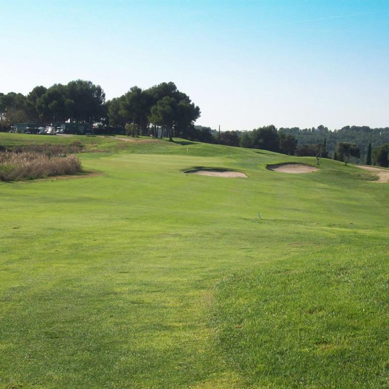 Panorama eines weitläufigen, gepflegten Golfplatzes mit grüner Rasenfläche, Sandbunkern und einem Waldhintergrund unter blauem Himmel.