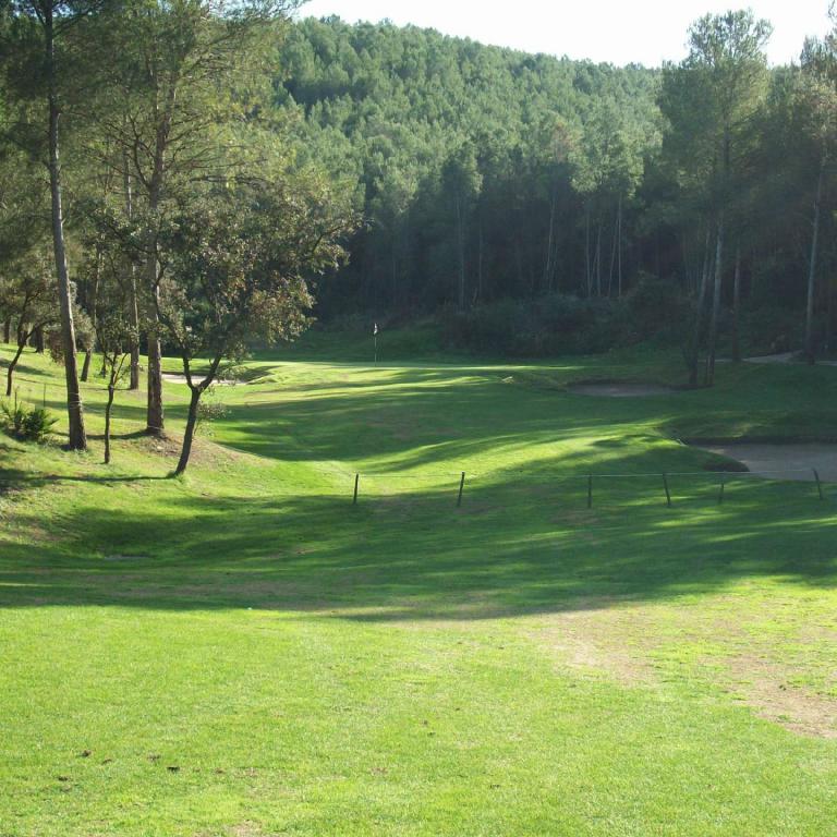 Ein Golfplatz mit grünem Rasen und Bäumen im Vordergrund, dahinter ein bewaldeter Hügel.