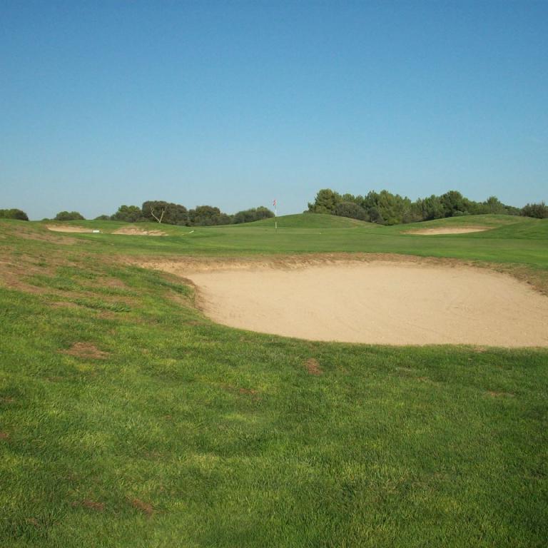 Ein Golfplatz mit einem großen Sandbunker im Vordergrund, umrahmt von grünem Gras und Bäumen unter blauem Himmel.