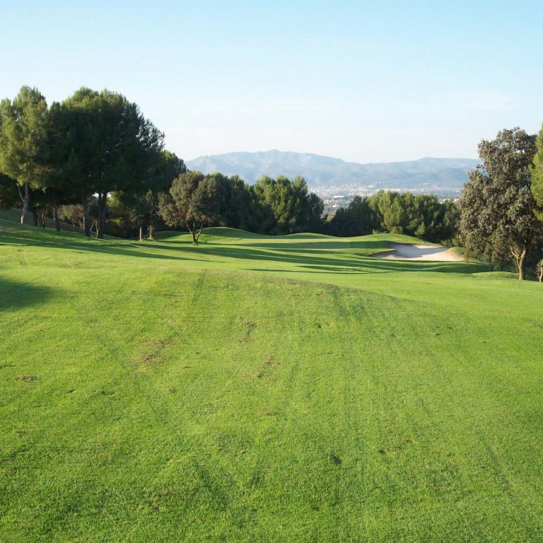 Ein weitläufiger, grüner Golfplatz, der sanft abfällt und im Hintergrund von dichtem Wald und einer Bergkulisse umrahmt wird.