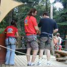 Mehrere Personen stehen auf einem Holzsteg, der von einer gespannten Seilkonstruktion überquert wird, mit Wald im Hintergrund.