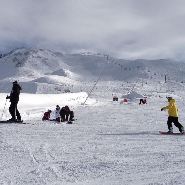 Weite Aufnahme eines Skigebiets mit präparierter Piste und mehreren Skifahrern unter bewölktem Himmel.