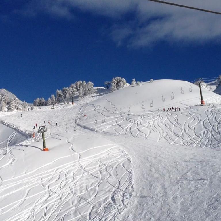 Weite Skipiste mit präpariertem Schnee, sichtbare Spuren und ein Seilbahnanlage unter blauem Himmel.