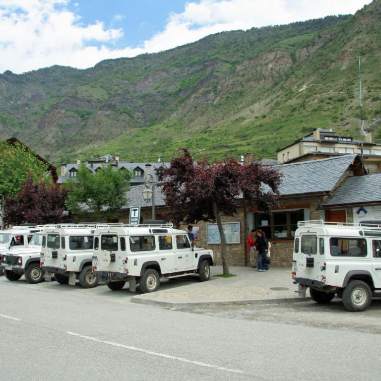 Ein Parkplatz mit mehreren weißen Geländewagen vor einem Dorf in einem Tal, umrahmt von steilen, grünen Berghängen.